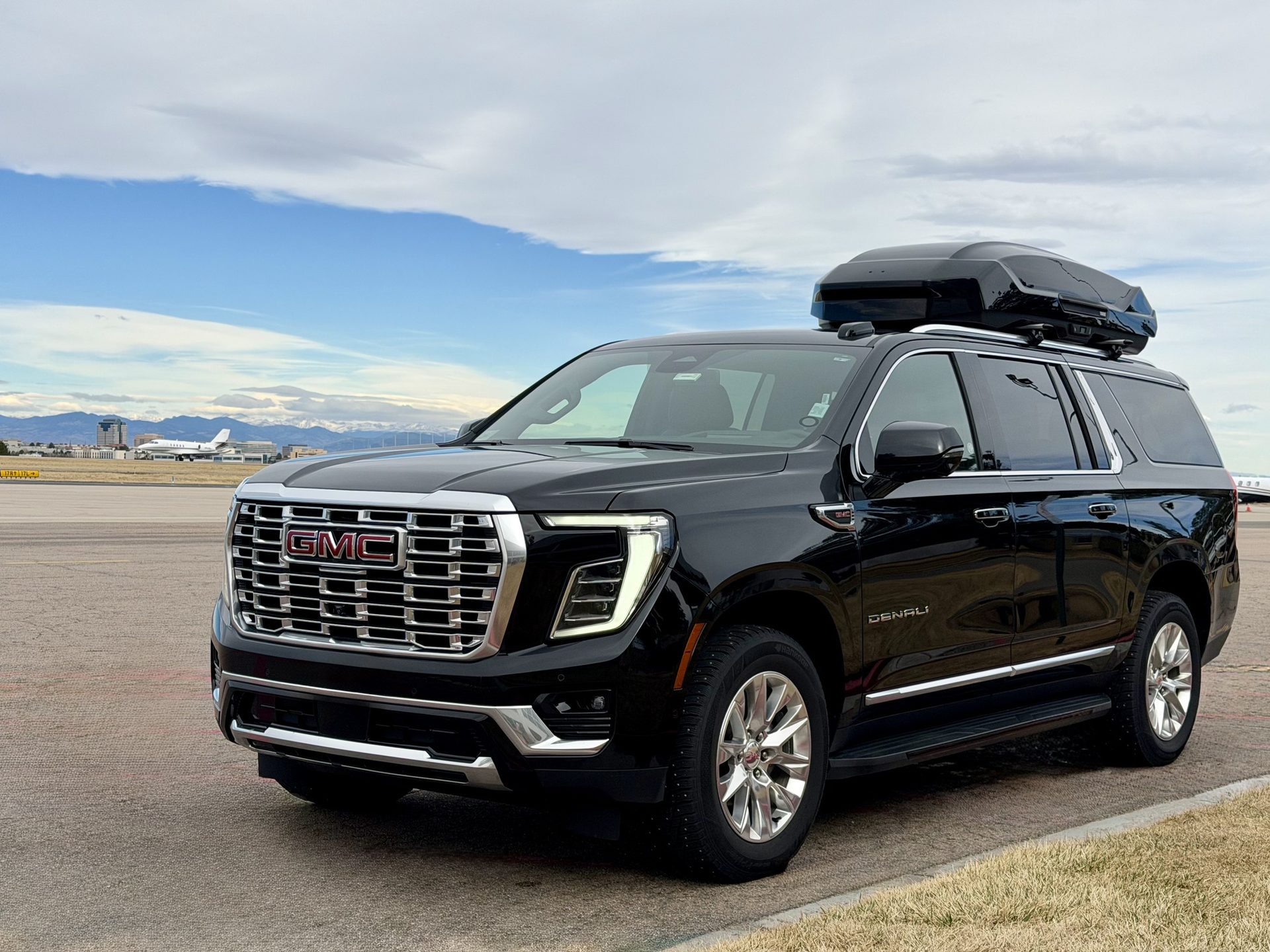 Black Blue Sky Limo GMC Yukon Denali XL with rooftop cargo box beside a private jet at a Denver-area airport.